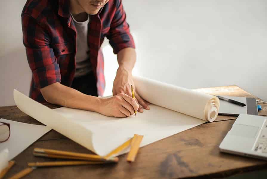 Homem de camisa xadrez desenhando em papel para projetos arquitetônicos. Na mesa, ferramentas para desenho técnico