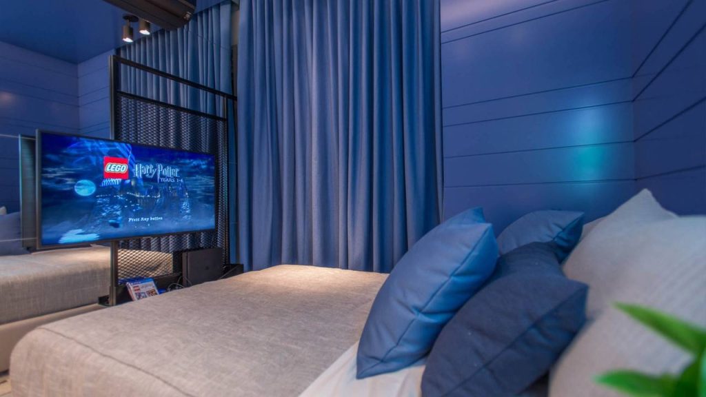 Quarto com decoração minimalista em tons de azul, em frente a cama uma Televisão ligada e ao lado uma parede com padrão geométrico