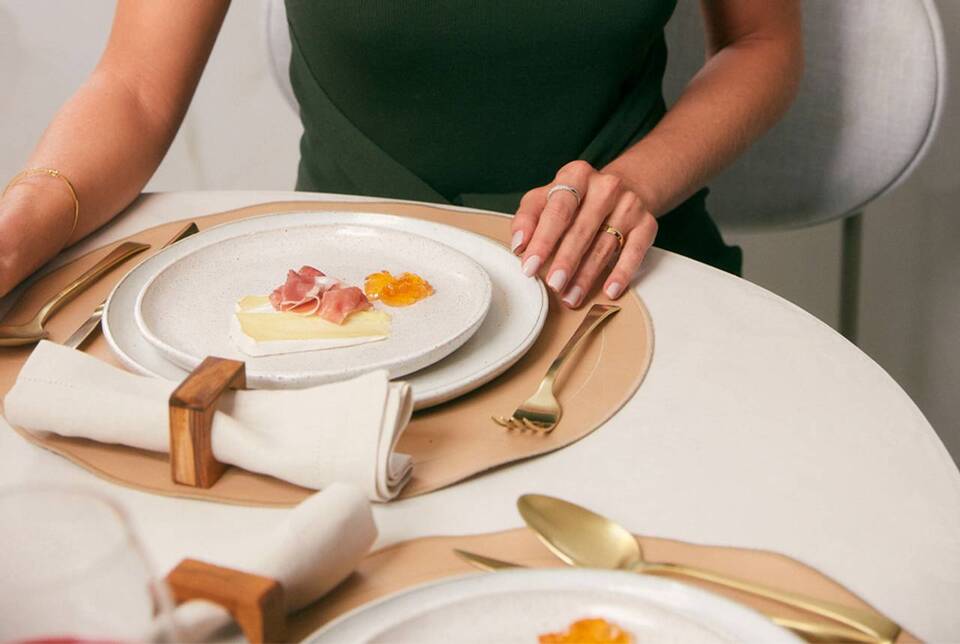 mulher sentada na mesa com os braços posturados segundo normas de etiqueta, um prato de de comida com queijo brie e presunto de parma