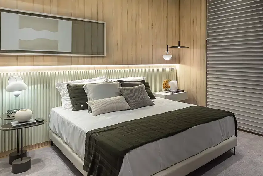 Quarto no estilo industrial com decoração de madeira clara e peças brancas, cama de casal com diversas almofadas e iluminação na cabeceira