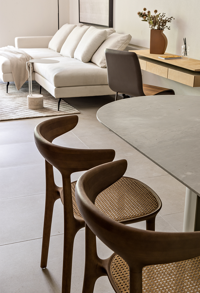 Uma sala de estar e jantar integrada com um sofá branco, mesa de jantar  com cadeiras de palha e madeira, e uma mesa de trabalho ao fundo, tudo em tons claros e neutros.